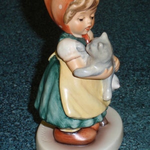 Puede incluir: Una figurita de porcelana de una ni&ntilde;a con un vestido rojo con lunares blancos y un gorro rojo que sostiene un gatito gris. La figurita est&aacute; de pie sobre una base blanca.