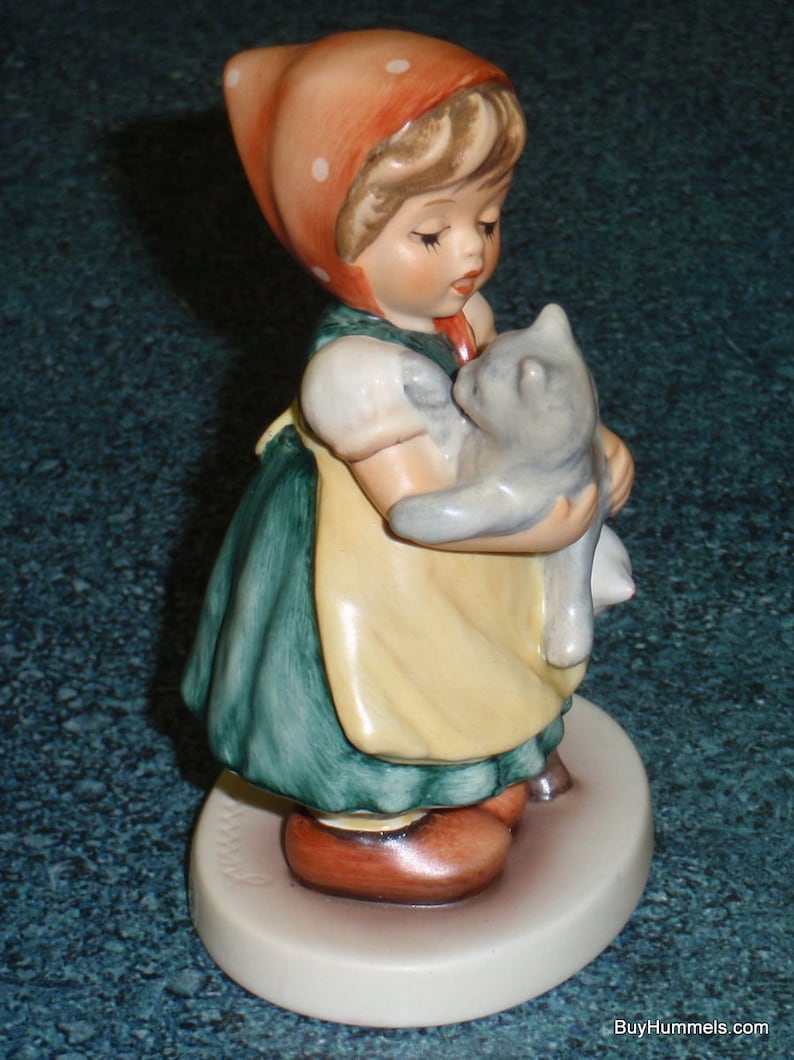 Puede incluir: Una figurita de porcelana de una ni&ntilde;a con un vestido rojo con lunares blancos y un gorro rojo que sostiene un gatito gris. La figurita est&aacute; de pie sobre una base blanca.