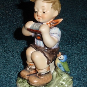 Può includere: Statuina in porcellana di un ragazzo seduto che suona l'armonica. Il ragazzo indossa pantaloncini marroni, stivali e una camicia bianca a righe blu. Un uccello blu e verde è seduto su un ciuffo d'erba accanto a lui.