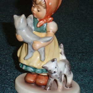 Puede incluir: Una figurita de una ni&ntilde;a con un vestido rojo con lunares blancos que sostiene un gato gris. Un gato blanco con manchas grises se sienta a su lado. La figurita est&aacute; sobre una base blanca.