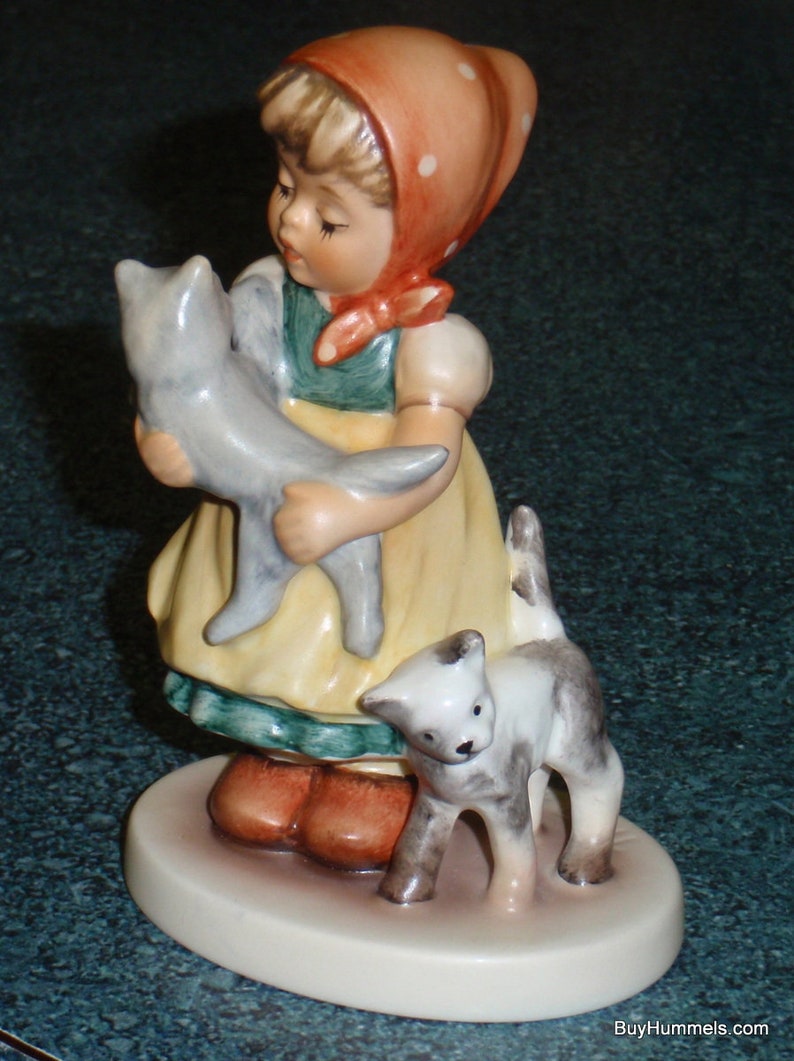 Puede incluir: Una figurita de una ni&ntilde;a con un vestido rojo con lunares blancos que sostiene un gato gris. Un gato blanco con manchas grises se sienta a su lado. La figurita est&aacute; sobre una base blanca.
