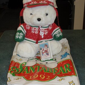 May include: A white plush bear wearing a red and white knit hat with earflaps and a matching red, white, and green zip-up sweater with snowflake patterns. A white musical note zipper pull is attached. A tag shows a Santa illustration with text.