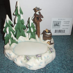 May include: A ceramic winter scene with a birdhouse, evergreen trees, and birds. The base is white with a light blue trim and purple flowers. The birdhouse is brown with a red roof, and the trees are green with white snow.