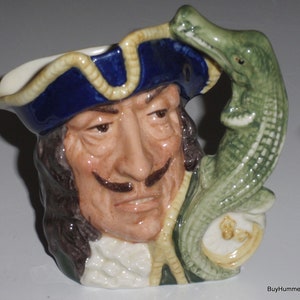 May include: A ceramic mug featuring a detailed depiction of a man with a mustache and a tricorn hat. The handle is a green alligator. The mug has a blue hat with a cream-colored trim and a cream-colored rim.