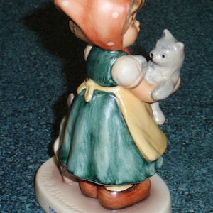 Puede incluir: Una figurita de una ni&ntilde;a con un vestido rojo con lunares blancos y un gorro rojo que sostiene un gatito gris. La figurita est&aacute; sobre una base blanca con las palabras "Katzenmama Tender Love" escritas en ella.