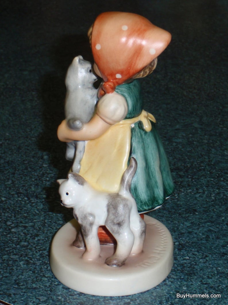 Puede incluir: Una figurita de una ni&ntilde;a con un vestido rojo y blanco de lunares y un gorro rojo que sostiene dos gatitos. La ni&ntilde;a est&aacute; de pie sobre una base blanca con la palabra "Hummel" escrita en ella.