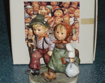 May include: A ceramic figurine of two children walking, a boy with a hat and a walking stick, and a girl with a red head covering and a bag. The figurine is on a round base with a green and white color scheme. The box behind the figurine has the text "M.J. Hummel The love lives on".