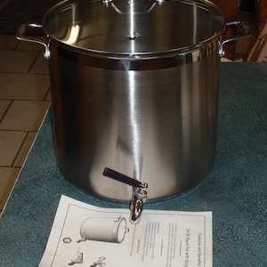 Puede incluir: Una olla grande de acero inoxidable con grifo, tapa de cristal y asas laterales. La olla está sobre una superficie azul con una hoja de papel impresa al lado. La olla está diseñada para cocinar y dispensar líquidos.