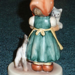 Puede incluir: Una figurita de una ni&ntilde;a con un vestido rojo con lunares blancos y un sombrero rojo que sostiene dos gatitos. La figurita est&aacute; sobre una base blanca con las palabras "Katzenmama Tender Love" escritas en ella.