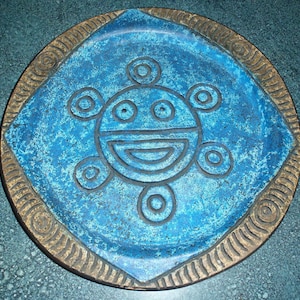 May include: A round, blue decorative plate with a central geometric design. The plate features a square center with a smiling face design and a textured, bronze-colored border. The plate is approximately 8 inches in diameter.