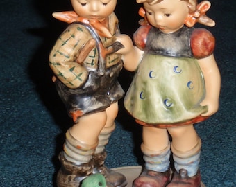 May include: A ceramic figurine depicting a boy and girl in traditional attire, standing on a base with a green frog. The boy wears a green hat with a feather, and the girl has red bows in her hair. The figurine is painted in muted tones.