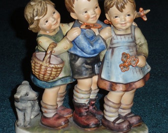 May include: A porcelain figurine of three children, a boy and two girls, standing together. The boy is wearing a blue shirt and brown pants, while the girls are wearing dresses. The girl on the left is holding a basket, and the girl on the right is holding a bouquet of flowers. A small dog is standing to the left of the children.