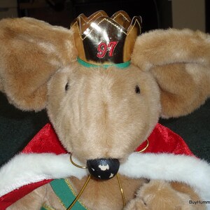 May include: A plush mouse wearing a gold crown with the number 97 in red, a red cape with white trim, and a green and gold sash. The mouse has large, fluffy ears and a black nose. The overall theme is a festive, possibly holiday-themed, collectible.