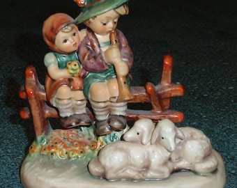 May include: A figurine of two children, a boy and a girl, sitting on a fence with three lambs lying at their feet. The boy is wearing a green hat and playing a flute. The girl is wearing a red hooded dress and holding a small bird. The figurine is made of porcelain and painted in bright colors.