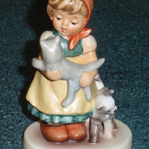 Puede incluir: Una figurita de porcelana de una ni&ntilde;a con un vestido rojo y blanco que sostiene un gato gris y un perro marr&oacute;n y blanco. La ni&ntilde;a lleva una diadema roja y blanca y un lazo rojo.