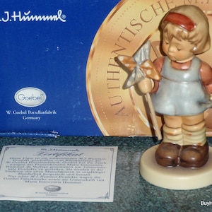 May include: A porcelain M.J. Hummel figurine of a child holding a pinwheel, standing on a round base. The child wears a blue dress, brown shoes, and a red headband. The set includes a blue branded box with gold text and a white paper certificate.