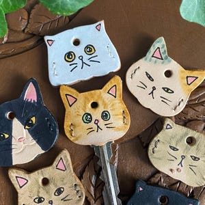 May include: A collection of handmade leather keychains shaped like cat faces. The keychains feature hand-drawn details, including eyes, whiskers, and stitched outlines. The color palette includes black, tan, orange, and white, with a key inserted into one of the keychains.