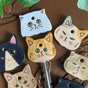 Puede incluir: Un conjunto de nueve llaveros de cuero con forma de gato y caras pintadas. Los llaveros son de varios colores, incluyendo negro, blanco, naranja y marrón. Cada llavero tiene una expresión diferente, algunos gatos parecen juguetones y otros parecen más serios.