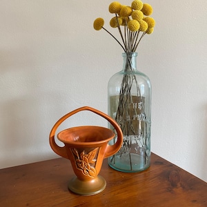 May include: A brown ceramic vase with a handle in the shape of a basket. The vase has a floral design and is sitting on a wooden table. A clear glass bottle with yellow dried flowers is in the background.