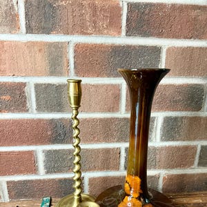 May include: Three brass candle holders, one tall and twisted, one short and simple, and one with a wide base. The candle holders are on a wooden surface in front of a brick wall. A brown ceramic vase with a floral design is in the background.