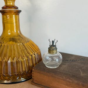 May include: A clear glass bottle with a metal top and a small wick. The bottle is sitting on a wooden box with the word "Townsends" printed on it. The bottle is next to a large amber glass decanter.