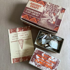 May include: An open box set with the text "ANCIENT CRAFT ARROWHEAD FLINT CHIP KIT" and a box of flint chips. A booklet with the text "ANCIENT CRAFT FLINT CHIP KIT MAKE YOUR OWN ARROWHEADS" is also visible. The set is designed for educational purposes.