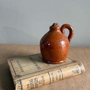 May include: A small, brown ceramic jug with a handle and a cork stopper. The jug is sitting on top of an antique book titled "The Mudfog Papers" by Dickens. The book has a beige cover with black text.