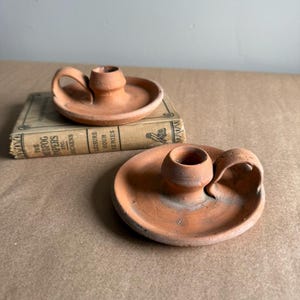 May include: Two terracotta candle holders with a circular base and a handle. The candle holders are a warm reddish-brown color and have a small cup to hold a candle. One is resting on a vintage book.