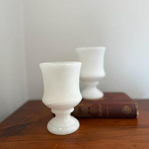 May include: Two white alabaster goblets with a classic chalice design. The goblets are displayed on a wooden surface, with one resting in front of the other. The goblets have a smooth, polished finish and a sturdy base.