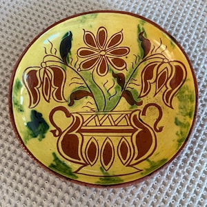 May include: A round, hand-painted ceramic plate with a yellow background and a brown floral design. The plate features a brown vase with flowers and leaves, and a green and brown border. The plate is approximately 8 inches in diameter.