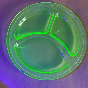 May include: A vintage, divided plate made of green glass. The plate has three sections and a raised rim. The glass glows brightly under a black light, showcasing its unique color and design. A classic piece for serving.