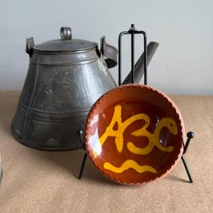 May include: A small, round, terracotta-colored bowl with the letters "ABC" in yellow paint, displayed on a black metal stand. A vintage metal kettle is in the background. The bowl has a wavy yellow design at the bottom.