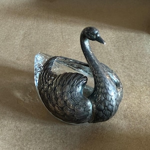 May include: A decorative swan-shaped dish with a silver-toned body and a clear glass bowl. The swan has a curved neck and detailed feathering. The dish is likely used for serving or display.