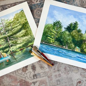 May include: Two watercolor paintings of a lakeside scene with lush green trees, a blue sky, and a house. One painting shows a dock with chairs. Paintbrushes are resting on the corner of the paintings. The artwork is displayed on a patterned surface.