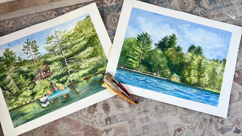 May include: Two watercolor paintings of a lakeside scene with lush green trees, a blue sky, and a house. One painting shows a dock with chairs. Paintbrushes are resting on the corner of the paintings. The artwork is displayed on a patterned surface.