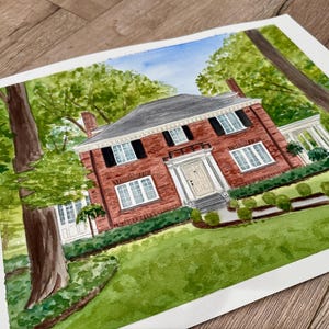 May include: Watercolor painting of a two-story brick house with a gray roof, black shutters, and white trim. The house is surrounded by green trees and a well-manicured lawn. The artwork is on white paper.