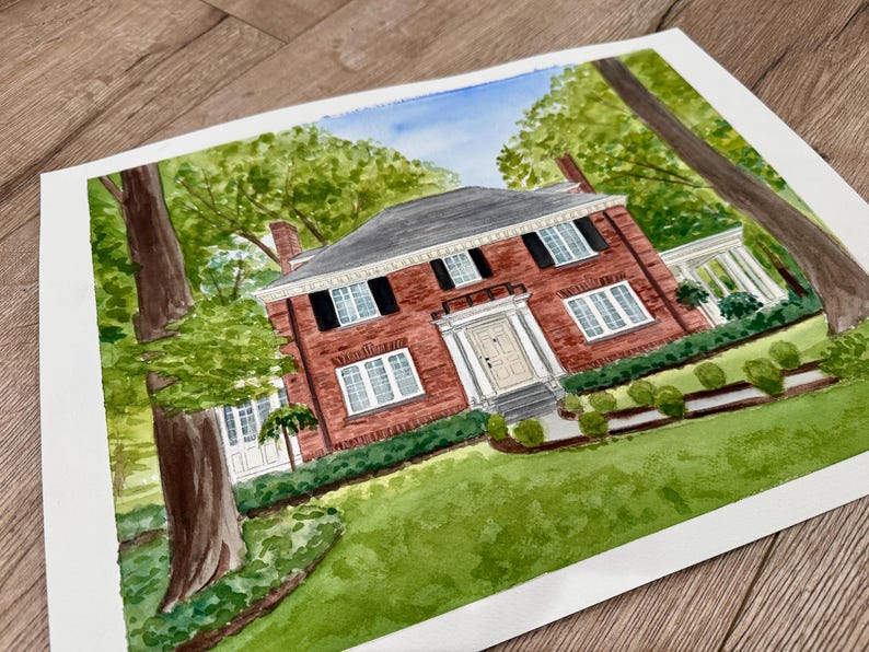 May include: Watercolor painting of a two-story brick house with a gray roof, black shutters, and white trim. The house is surrounded by green trees and a well-manicured lawn. The artwork is on white paper.