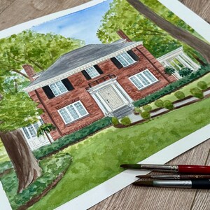 May include: Watercolor painting of a red brick house with white trim, black shutters, and a dark gray roof. The house is surrounded by green grass, trees, and bushes. Paintbrushes are in the foreground.