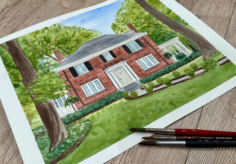 May include: Watercolor painting of a red brick house with white trim, black shutters, and a dark gray roof. The house is surrounded by green grass, trees, and bushes. Paintbrushes are in the foreground.
