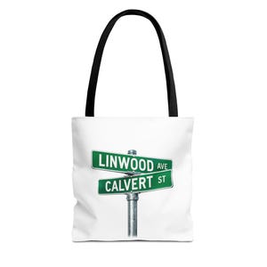 Könnte beinhalten: Weiße Tragetasche mit schwarzen Griffen. Sie zeigt ein grünes Straßenschild mit der Aufschrift "LINWOOD AVE" und "CALVERT ST". Die Tasche besteht aus strapazierfähigem Material und eignet sich zum Tragen verschiedener Gegenstände.