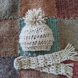 Op de afbeelding: Een beige gebreide muts met een pompon bovenop en een bijpassend paar gebreide vingerloze handschoenen. De muts en handschoenen liggen op een geweven stoffen achtergrond.
