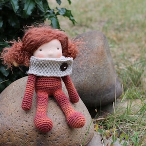 Puede incluir: Muñeca de ganchillo hecha a mano con pelo rojo y una bufanda gris. La muñeca está sentada en una gran roca. El cuerpo y las extremidades de la muñeca son de tonos rojos y naranjas. La muñeca tiene un botón en la bufanda.