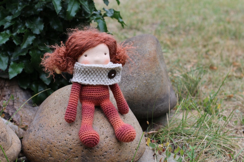 May include: A handmade crochet doll with red hair and a gray scarf. The doll is sitting on a large rock. The doll's body and limbs are shades of red and orange. The doll has a button on the scarf.