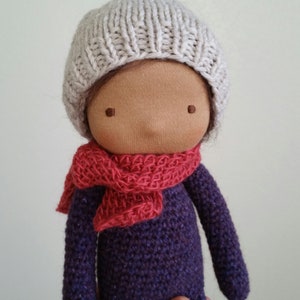 May include: A handmade doll with a light beige knitted hat and a red scarf. The doll is wearing a purple knitted sweater. The doll has a tan face with simple stitched features.