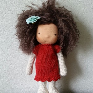 May include: A handmade doll with a tan face, brown curly hair, and a light blue flower. The doll wears a red knitted dress with a scalloped hem, white knitted arms, and white knitted legs. The doll is standing upright.