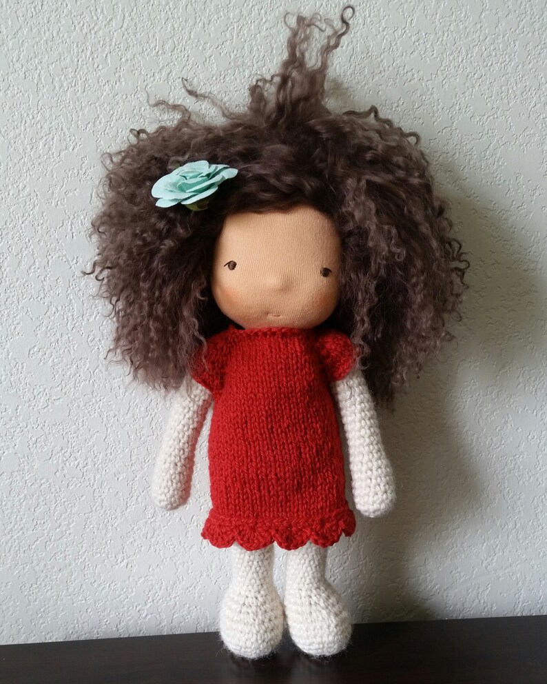 May include: A handmade doll with a tan face, brown curly hair, and a light blue flower. The doll wears a red knitted dress with a scalloped hem, white knitted arms, and white knitted legs. The doll is standing upright.