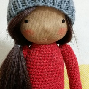 May include: A handmade doll with a light brown face, rosy cheeks, and dark brown hair styled in a ponytail. The doll wears a gray knit cap and a red crocheted sweater. The doll's eyes are small black dots.