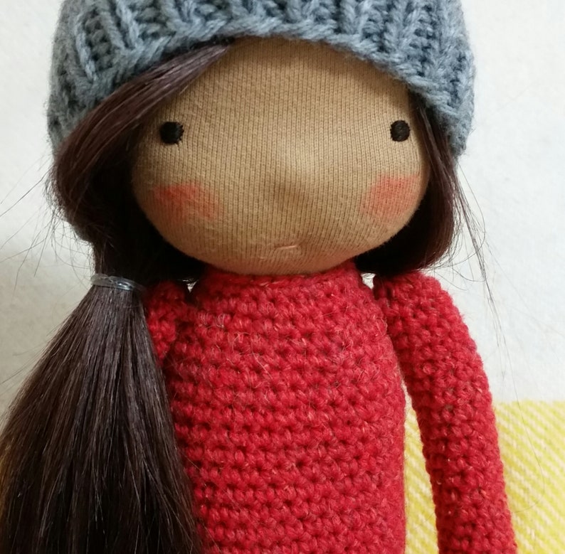 May include: A handmade doll with a light brown face, rosy cheeks, and dark brown hair styled in a ponytail. The doll wears a gray knit cap and a red crocheted sweater. The doll's eyes are small black dots.