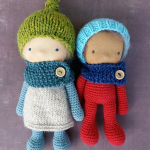 May include: Two handmade dolls with fabric faces, wearing knitted hats and scarves. One doll has a green hat and a gray and blue outfit. The other doll is dressed in red with a blue hat and scarf. Each doll has a button closure on the scarf.