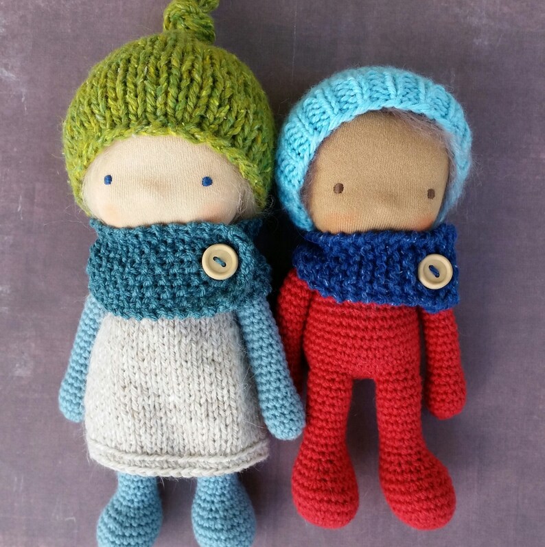 May include: Two handmade dolls with fabric faces, wearing knitted hats and scarves. One doll has a green hat and a gray and blue outfit. The other doll is dressed in red with a blue hat and scarf. Each doll has a button closure on the scarf.